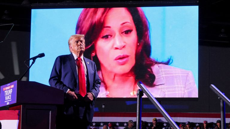 Republican presidential nominee and former US.President Donald Trump looks on as Democratic presidential nominee and US Vice President Kamala Harris' face appears as a video plays on a screen, during a rally at Huntington Place in Detroit, Michigan