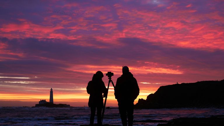 Lidé fotí barevnou oblohu před východem slunce na majáku St Mary's ve Whitley Bay na severovýchodním pobřeží Anglie. V některých částech Spojeného království byl předpovídán vítr o rychlosti až 75 mil za hodinu, protože období vánočních útěků nadále zaznamenává narušení způsobené špatným počasím. Datum snímku: pondělí 23. prosince 2024.