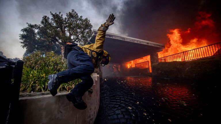 Hasič skáče přes plot při hašení požáru Palisades ve čtvrti Pacific Palisades v Los Angeles, úterý 7. ledna 2025. (AP Photo/Ethan Swope)